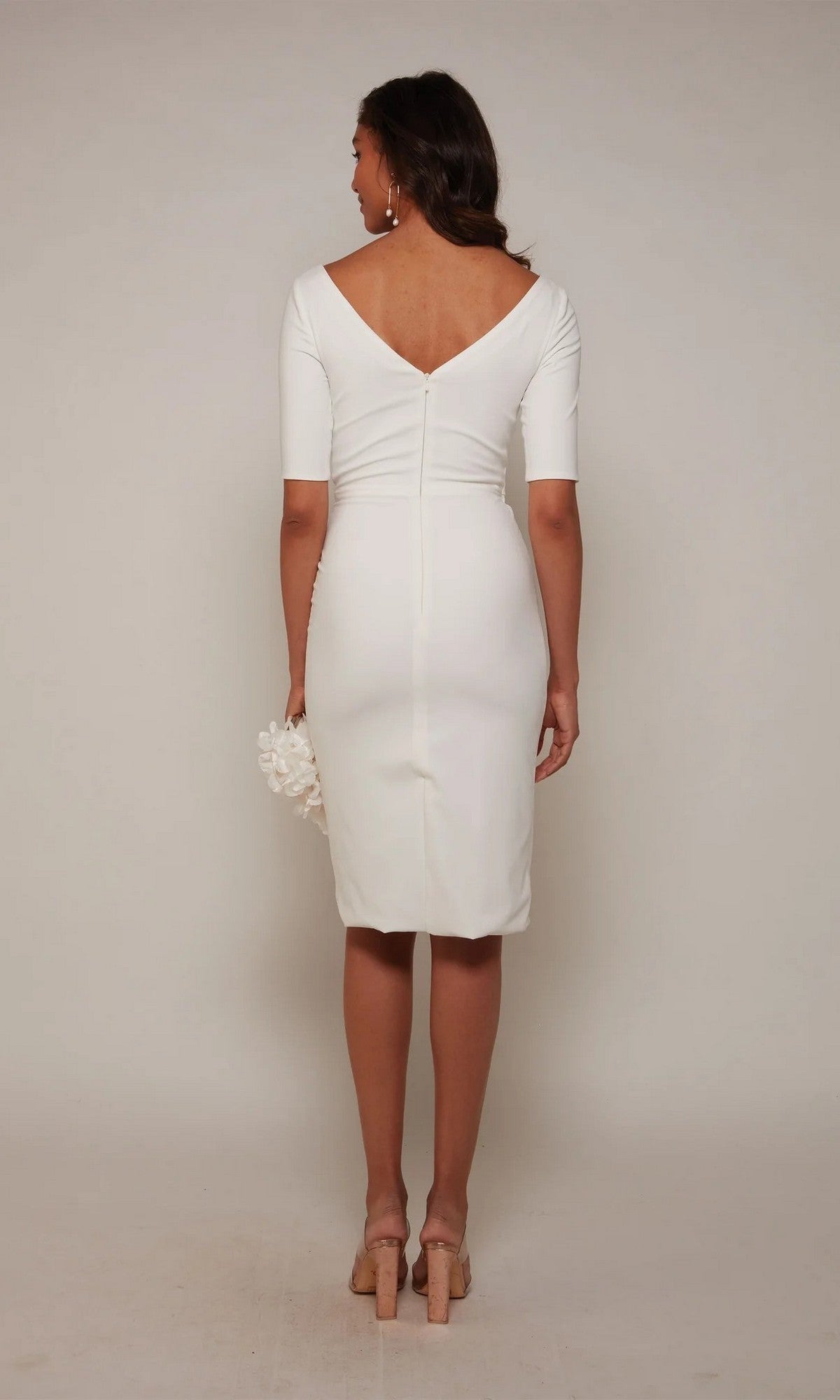 A woman faces away in the Alyce 70036 Knee Length Semi-Formal Dress, featuring a deep V-back and short sleeves. She holds a small white accessory and wears nude heels—ideal for cocktail parties, set against a plain background.